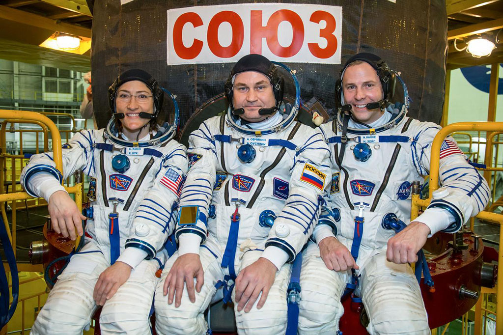 Expedition crew wearing spacesuits and sitting in from of Soyuz craft